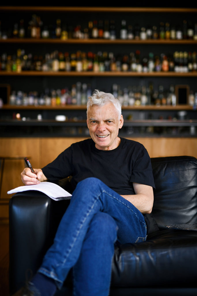 Graeme Simsion wearing blue jeans and black t-shirt, sitting on a black leather sofa, writing in a note book.